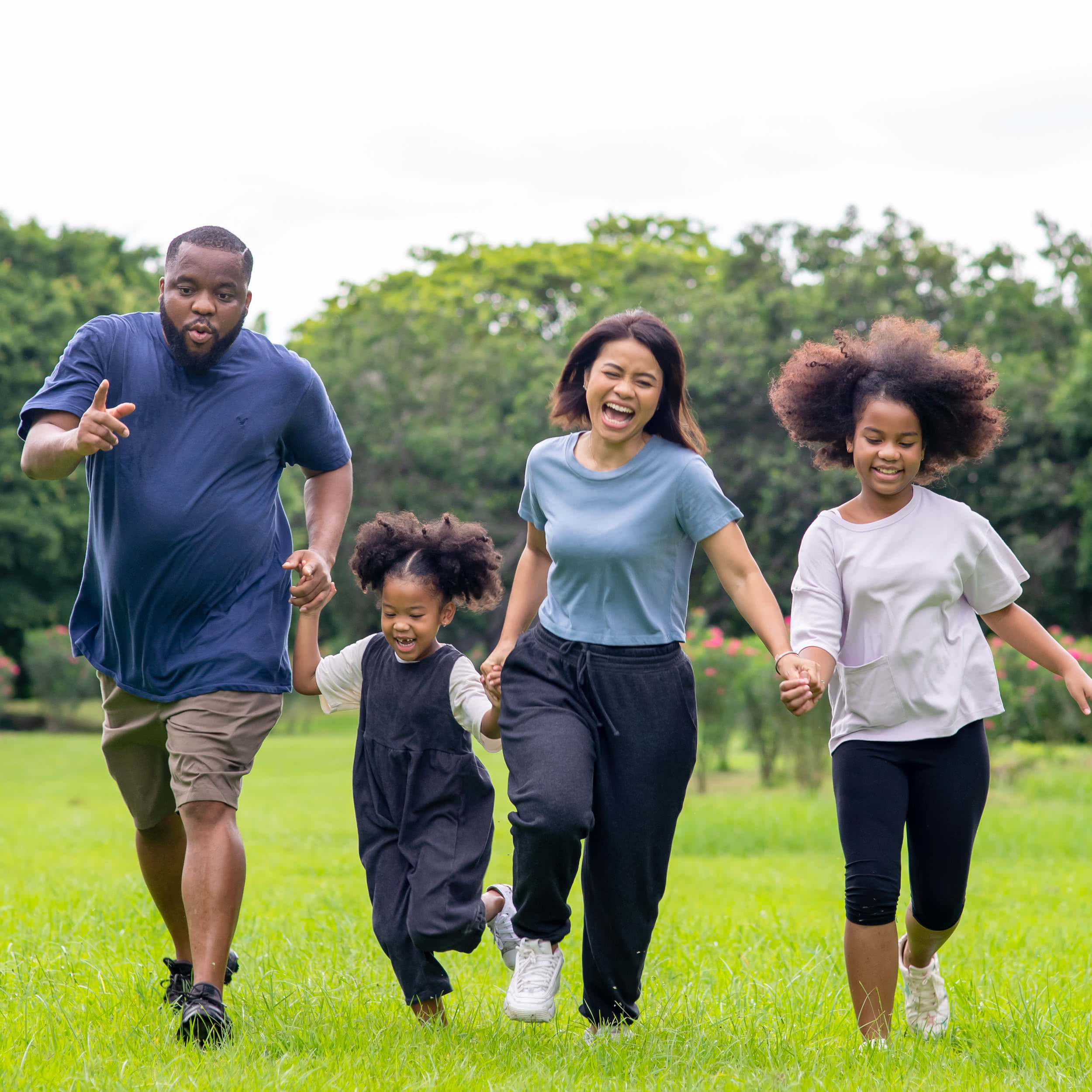 A young, happy family holds hands and runs together through a grassy field in a park, surrounded by trees