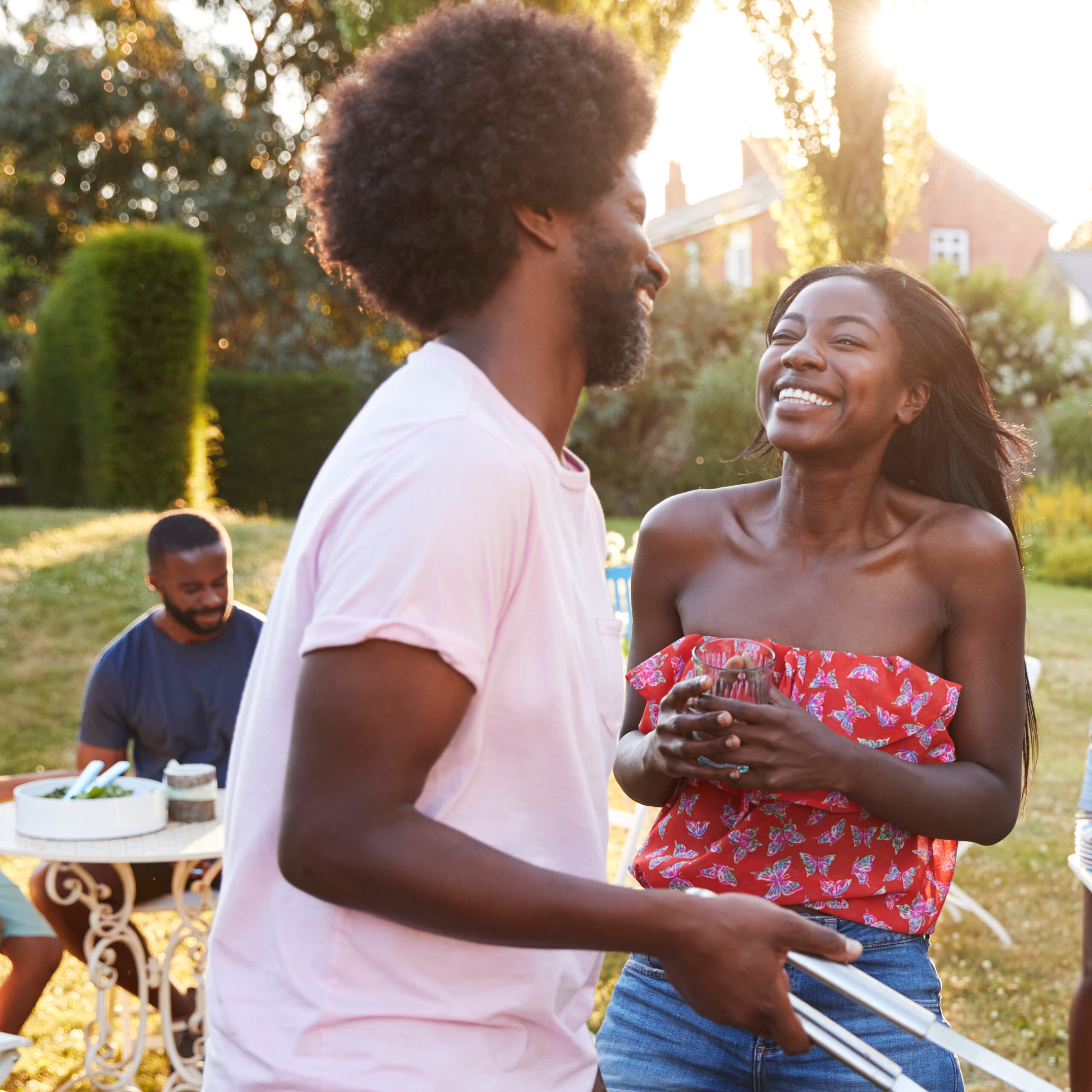 Couple laughing, barbecuing together outside as friend sits in the background at a table