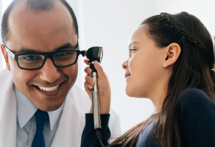 A young girl inspects a smiling doctor's ear with an otoscope